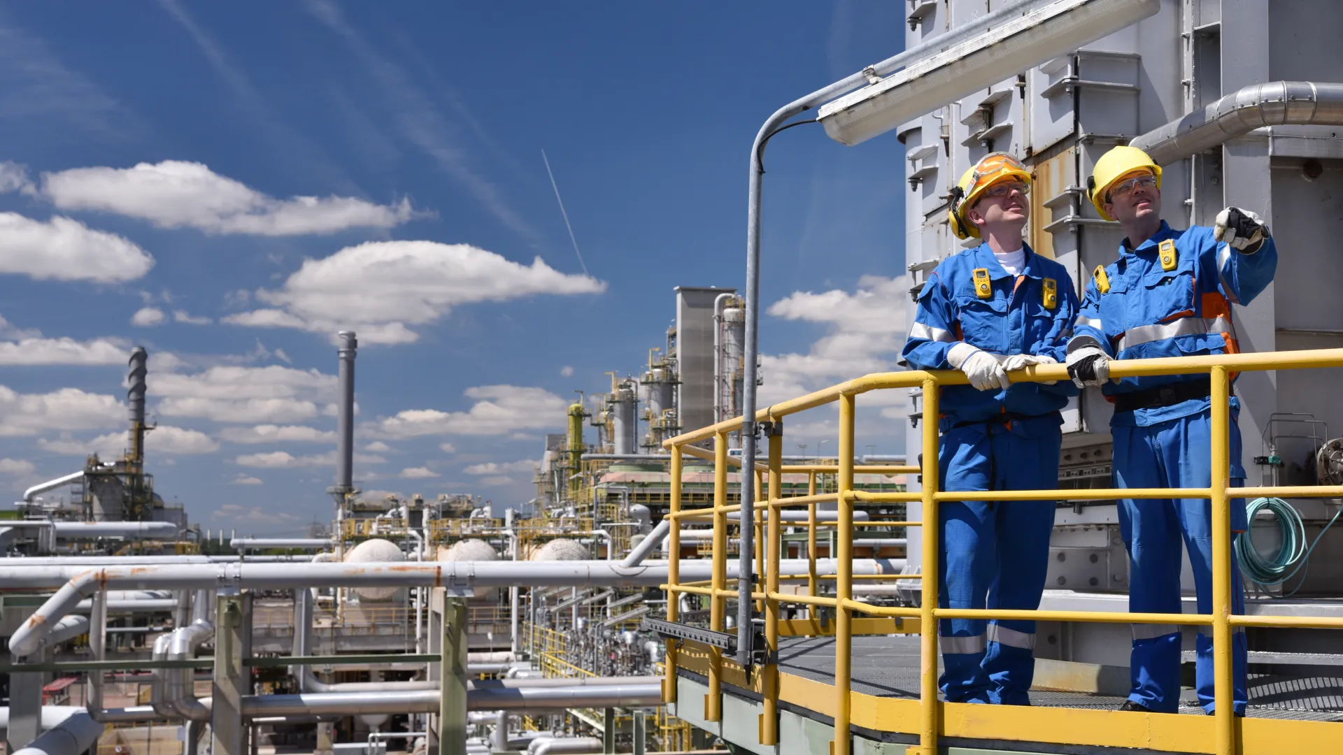 Blick von schräg oben auf Petrochemieanlage, rechts stehen diskutierend zwei Männer mit gelbem Helm und Blaumann auf einer Ballustrade
